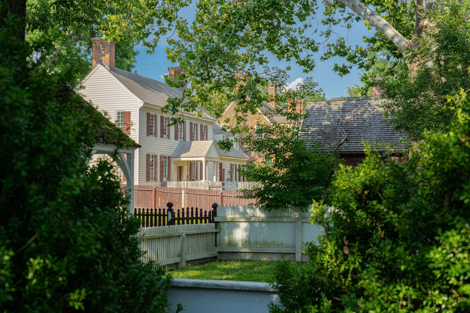 White colonial-era homes with brick chimneys and picket fencing behind mature trees