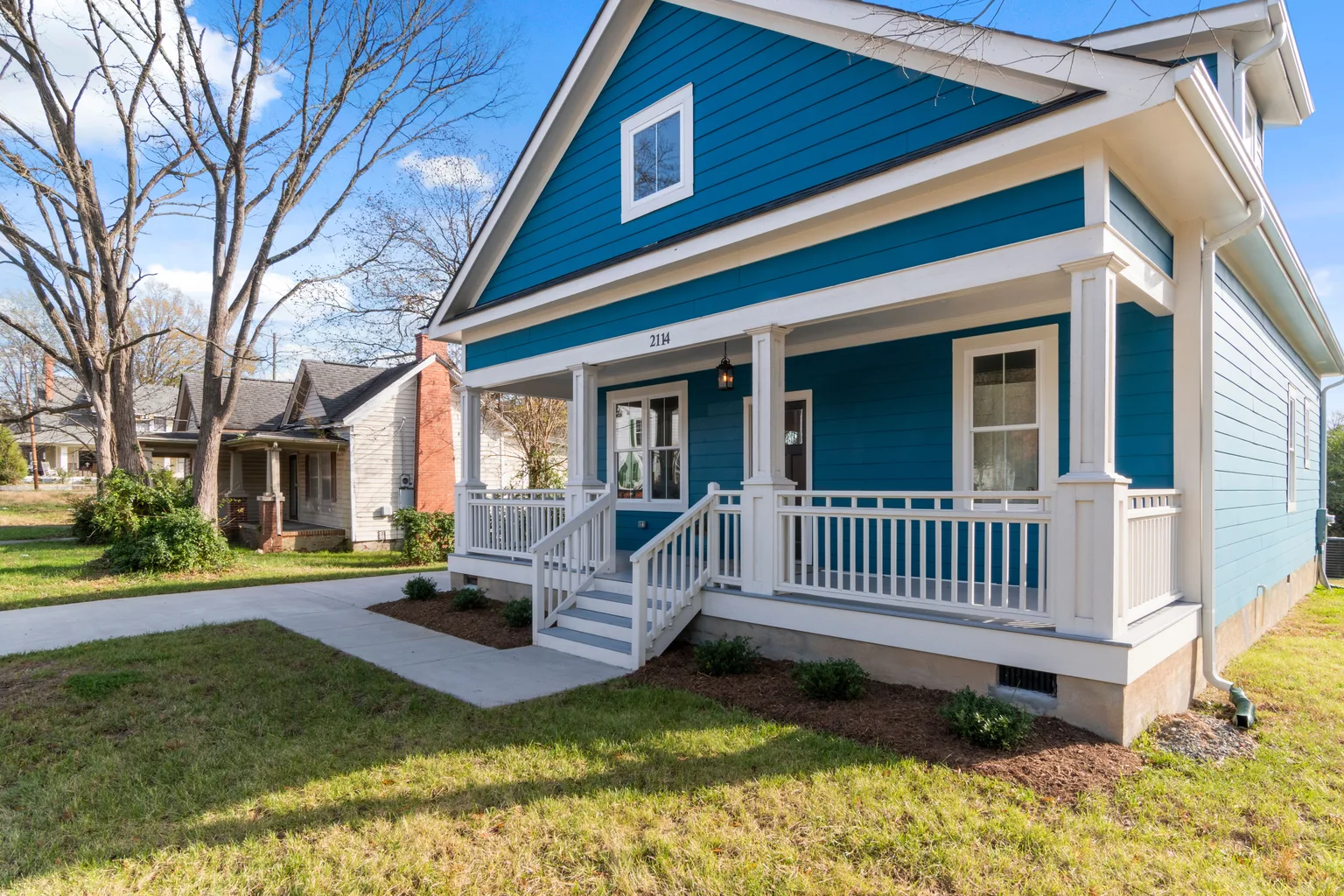 A blue bungalow with a covered front porch surrounded by mature trees on a clear spring day