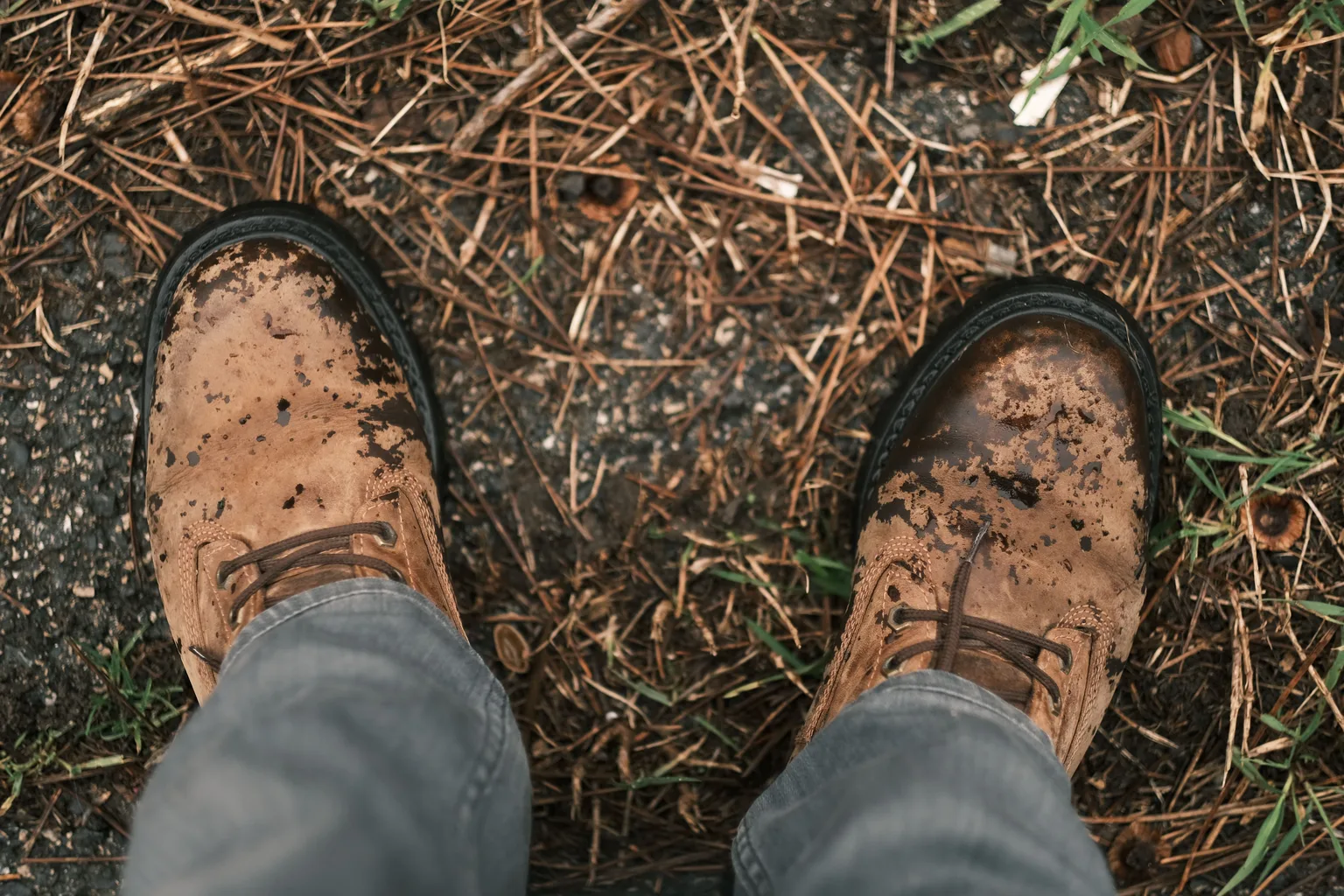 Worn brown leather boots caked with dried mud standing on a rough outdoor surface
